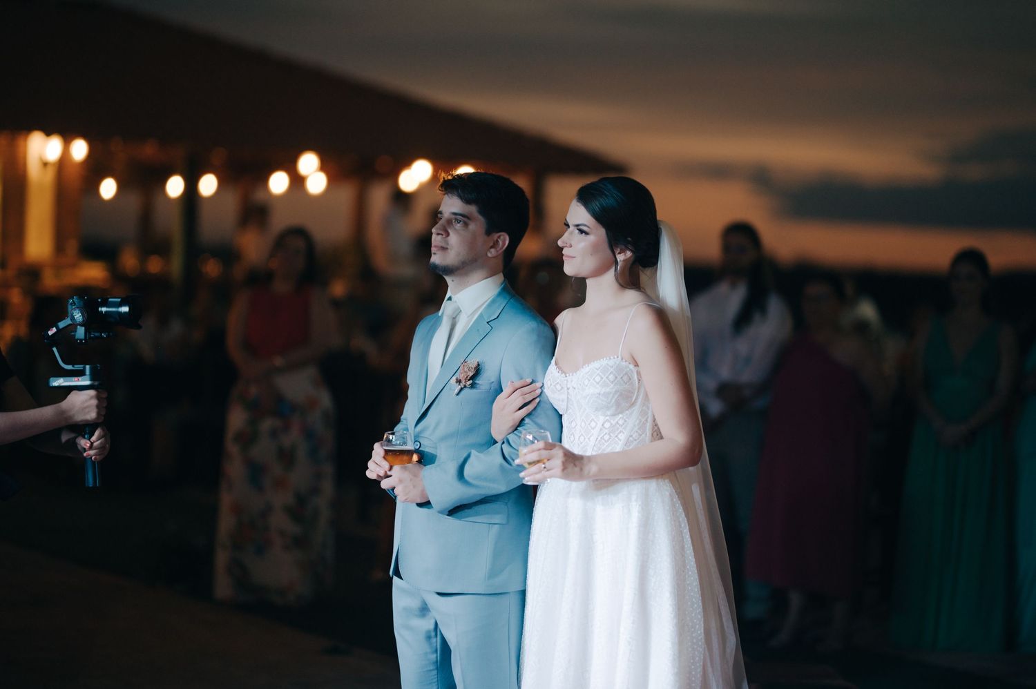 A couple stands side by side during an outdoor twilight celebration with bokeh lights.