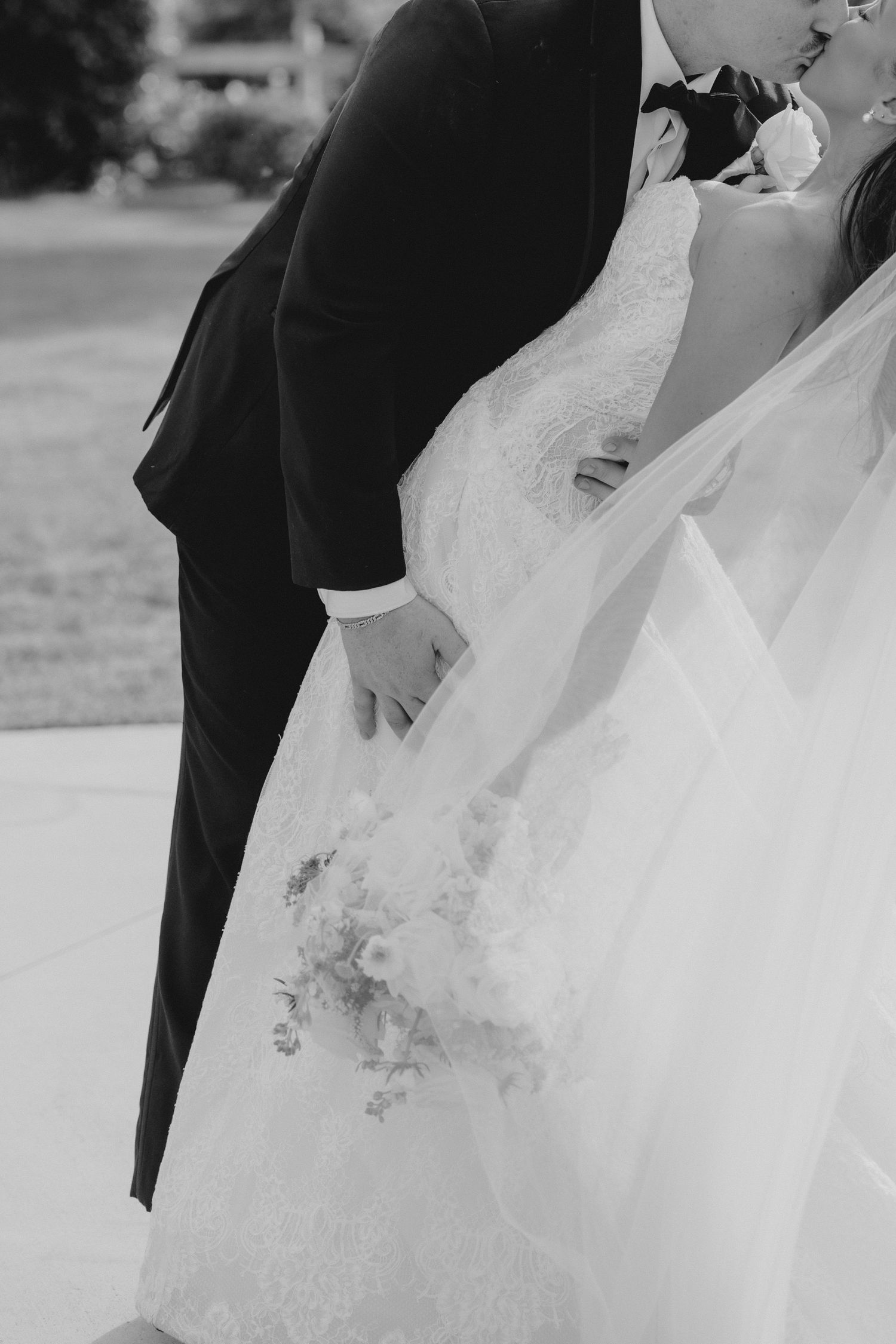 Black and white photo of bride and groom sharing a kiss, bride holding a floral bouquet in a lace wedding gown. Fresno Wedding Videographer
