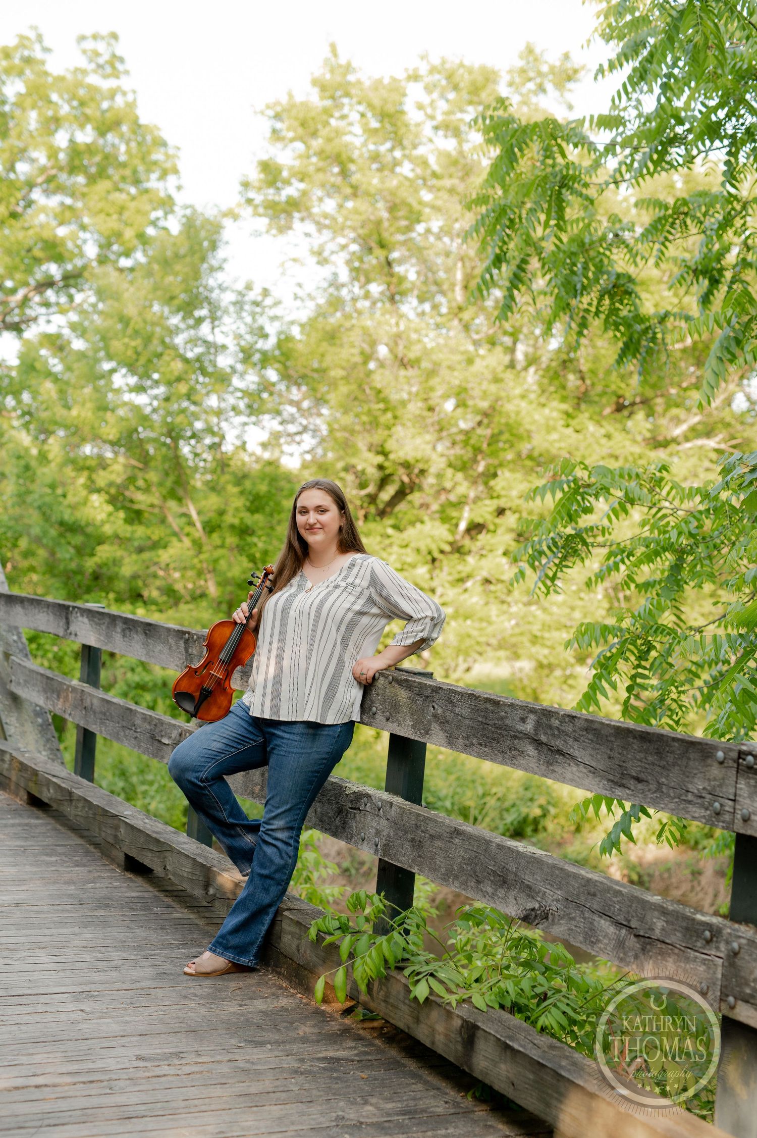 Musician with violin poses against wooden railing on bridge surrounded by trees.