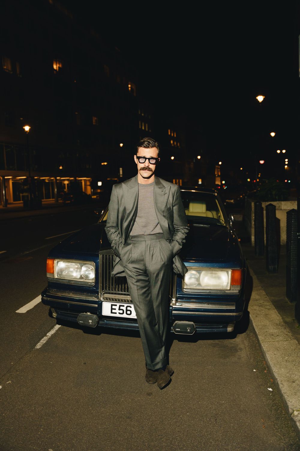 A person in a grey suit and glasses poses at night in front of a classic dark-coloured luxury car on a London street photographed with flash
