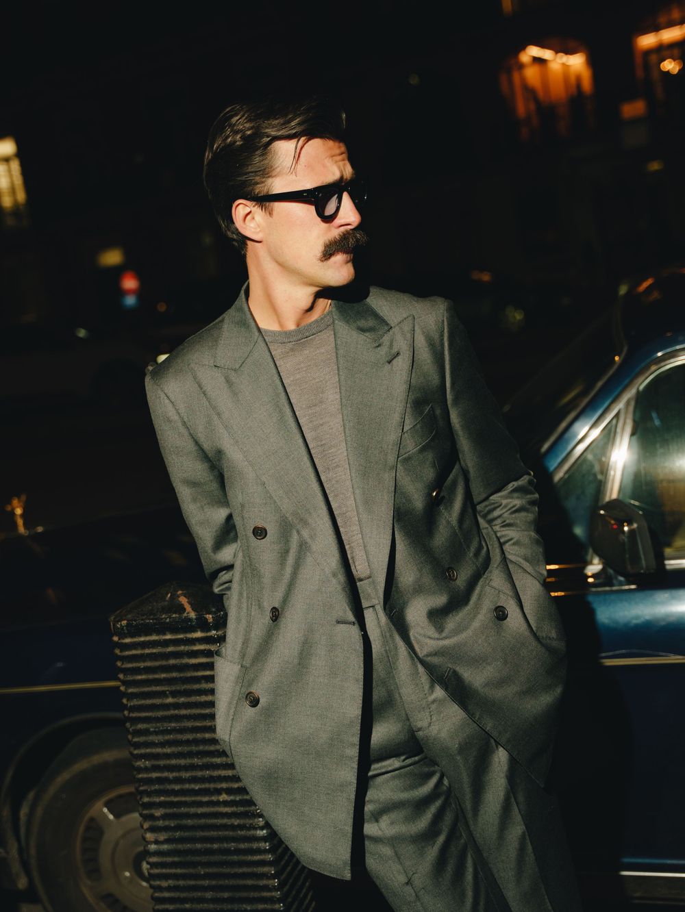 A person in sunglasses wearing a double-breasted gray suit with a casual t-shirt underneath poses on a dark street - flash photography