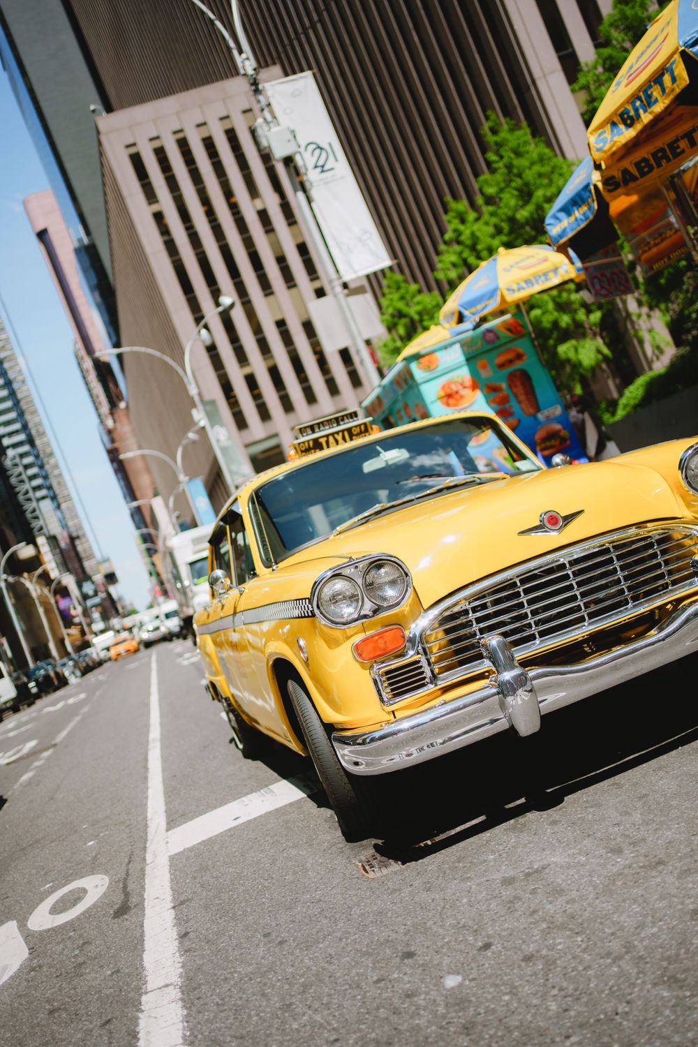 A vintage yellow Checker taxi cab parked on a city street with tall buildings in the background and trees along the sidewalk.