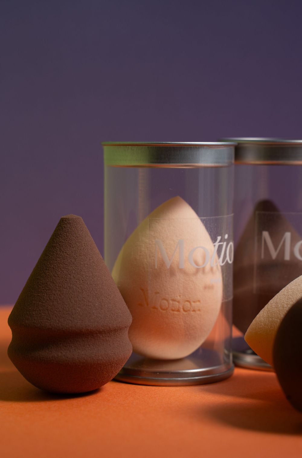 Conical and egg-shaped makeup sponges displayed with Motion branded containers on orange surface.