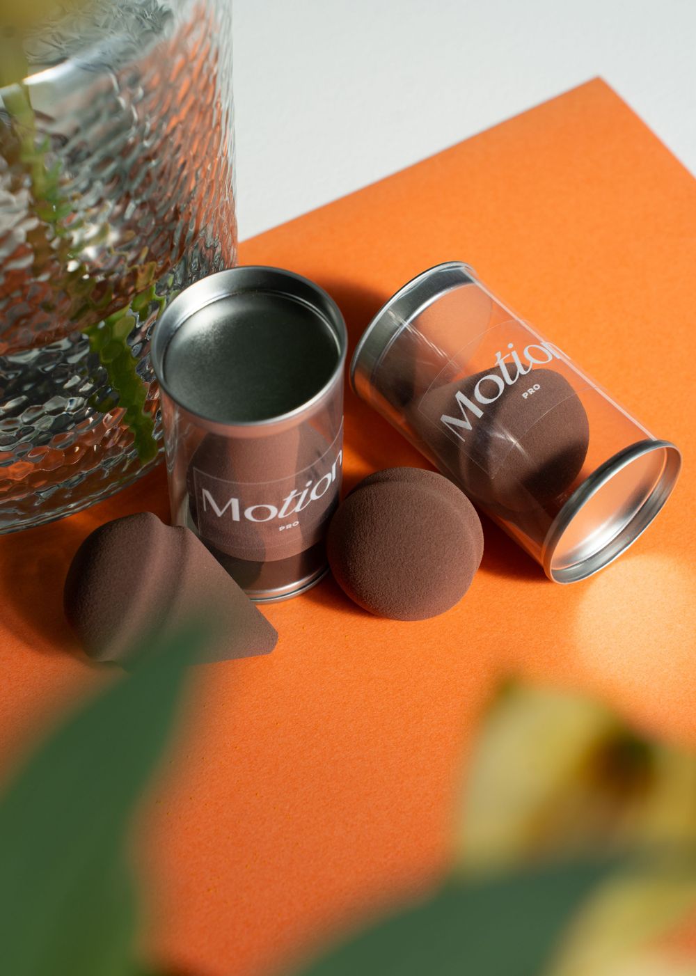 Motion branded containers with brown makeup sponges displayed on orange surface with plants.