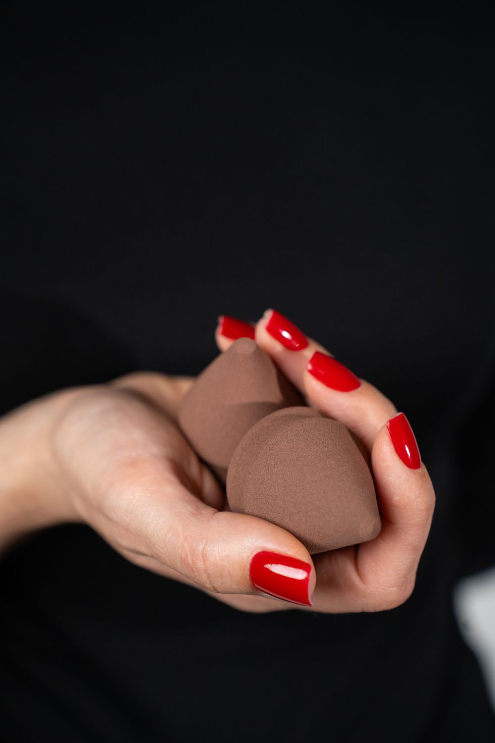 Red-manicured hands holding brown makeup sponge against dark background.