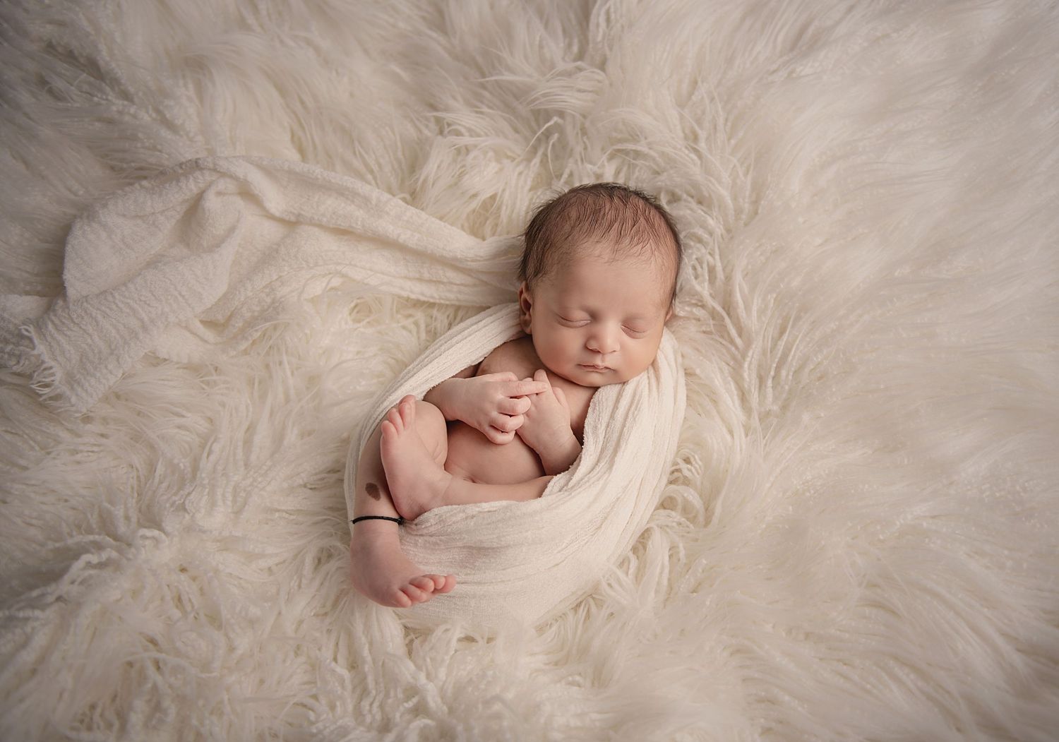 Sleeping newborn wrapped in cream fabric nestled on white fur backdrop.