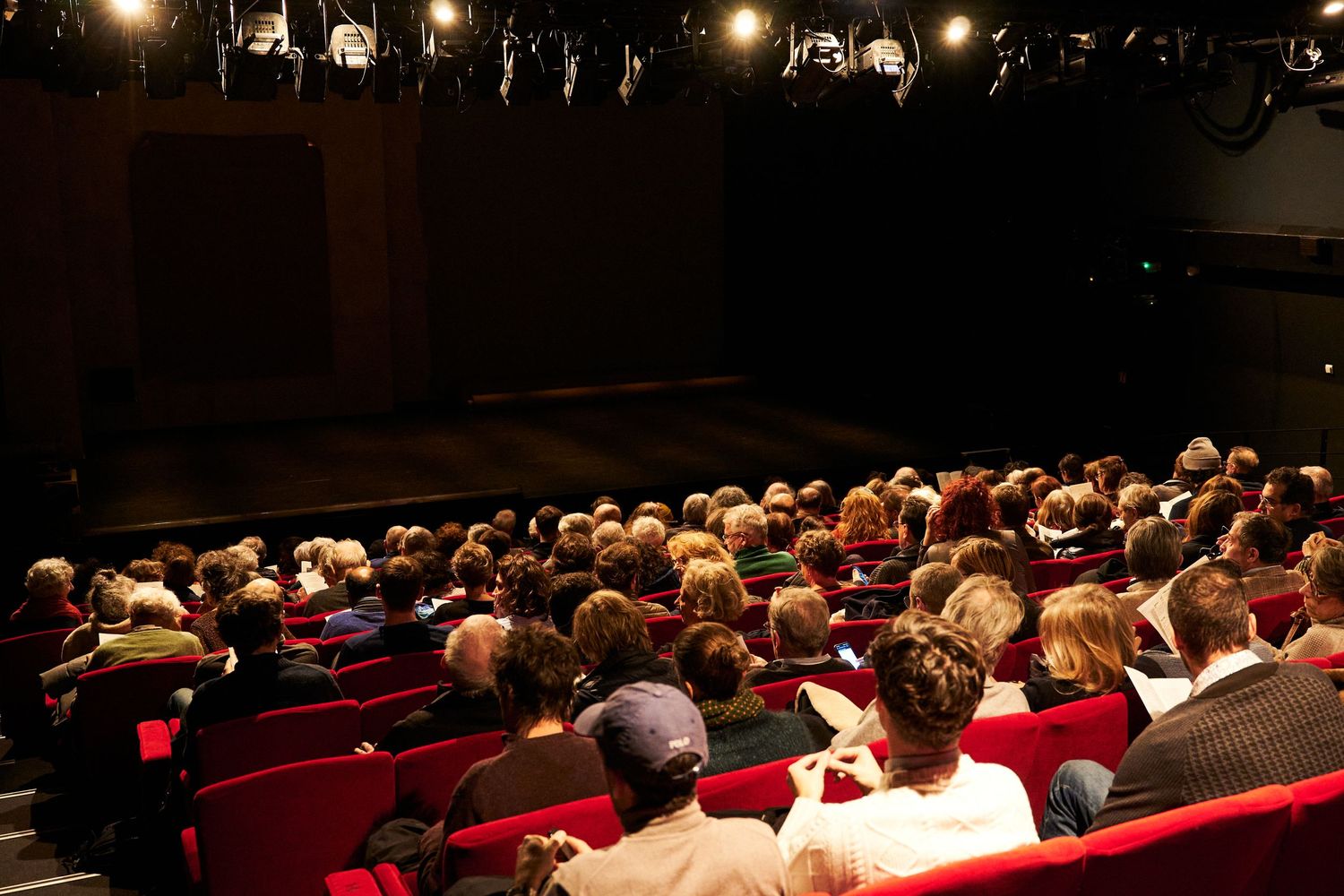 Public de théâtre assis sur des chaises rouges regardant une représentation.