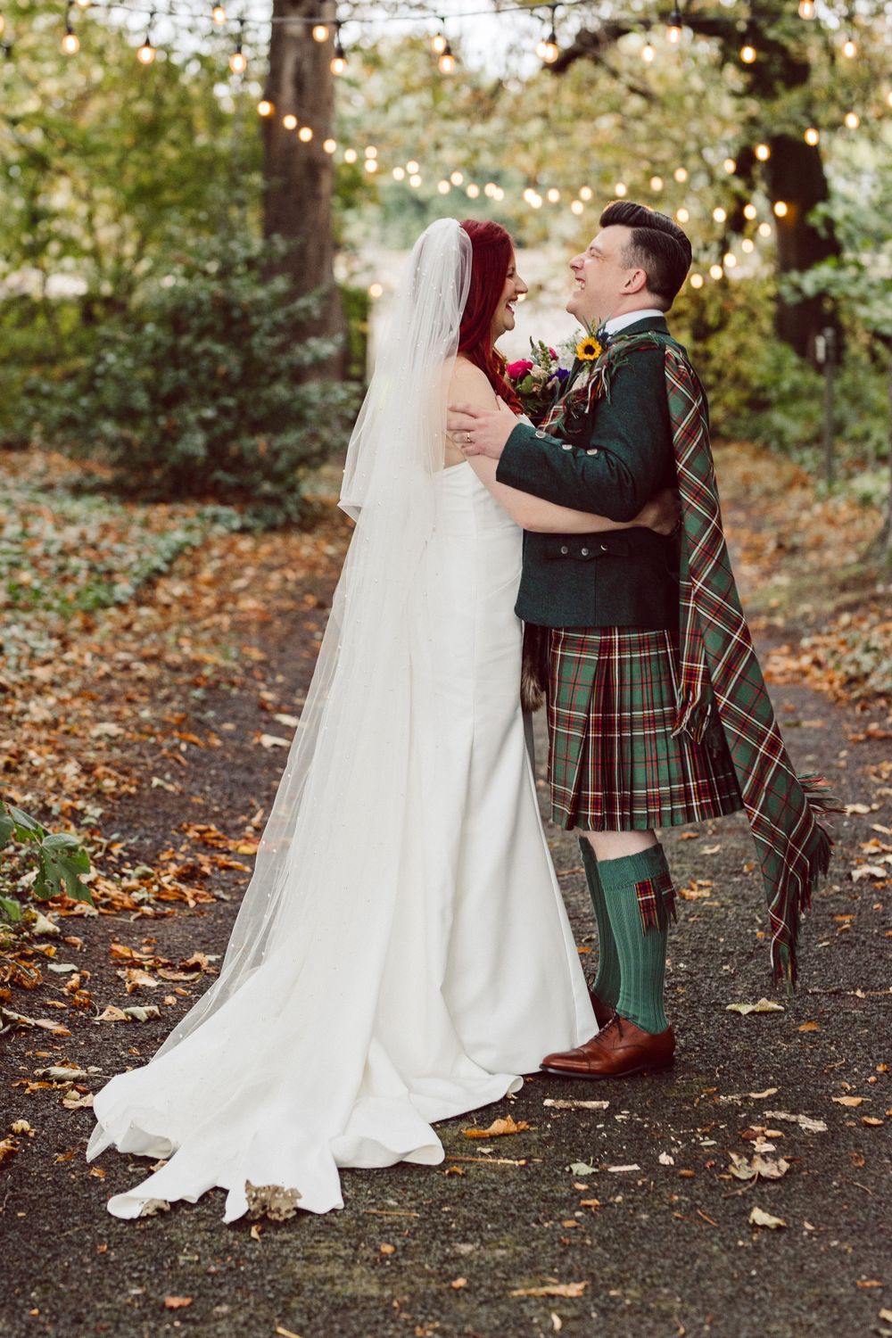 Romantic wedding couple embracing on autumn path with string lights and Scottish attire.