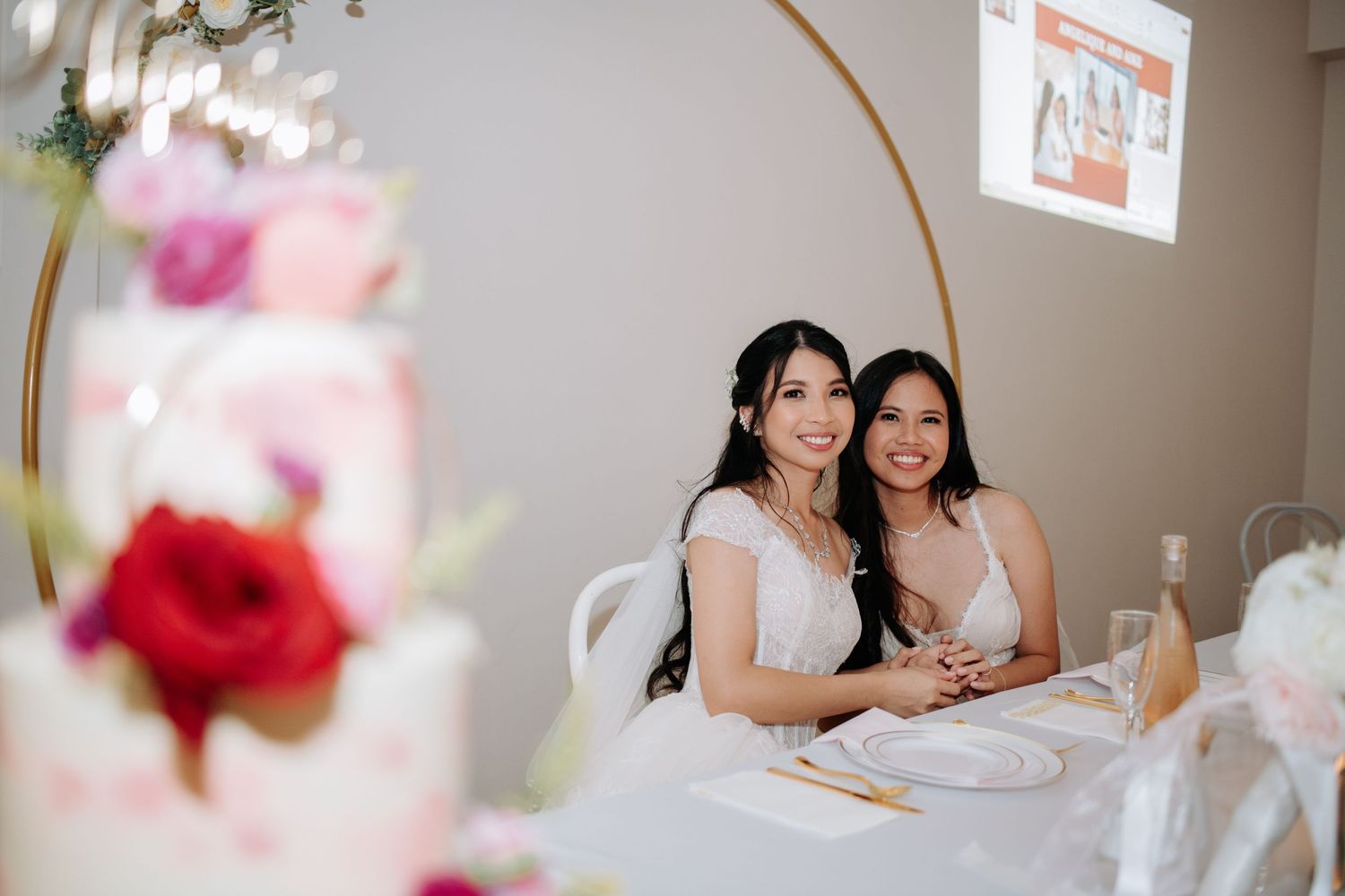 Two people in white dresses seated at decorated wedding reception table with floral centerpiece.