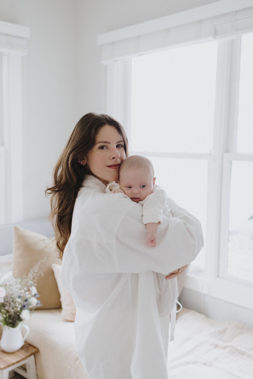 Mother in white dress holding and kissing baby near bright window.