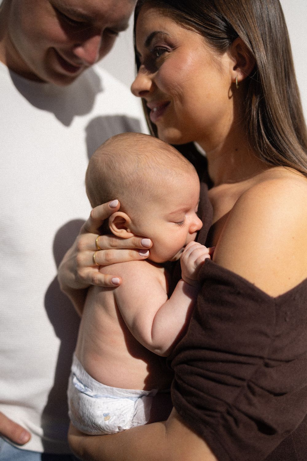 Mother in brown sweater holding infant against bright white wall.