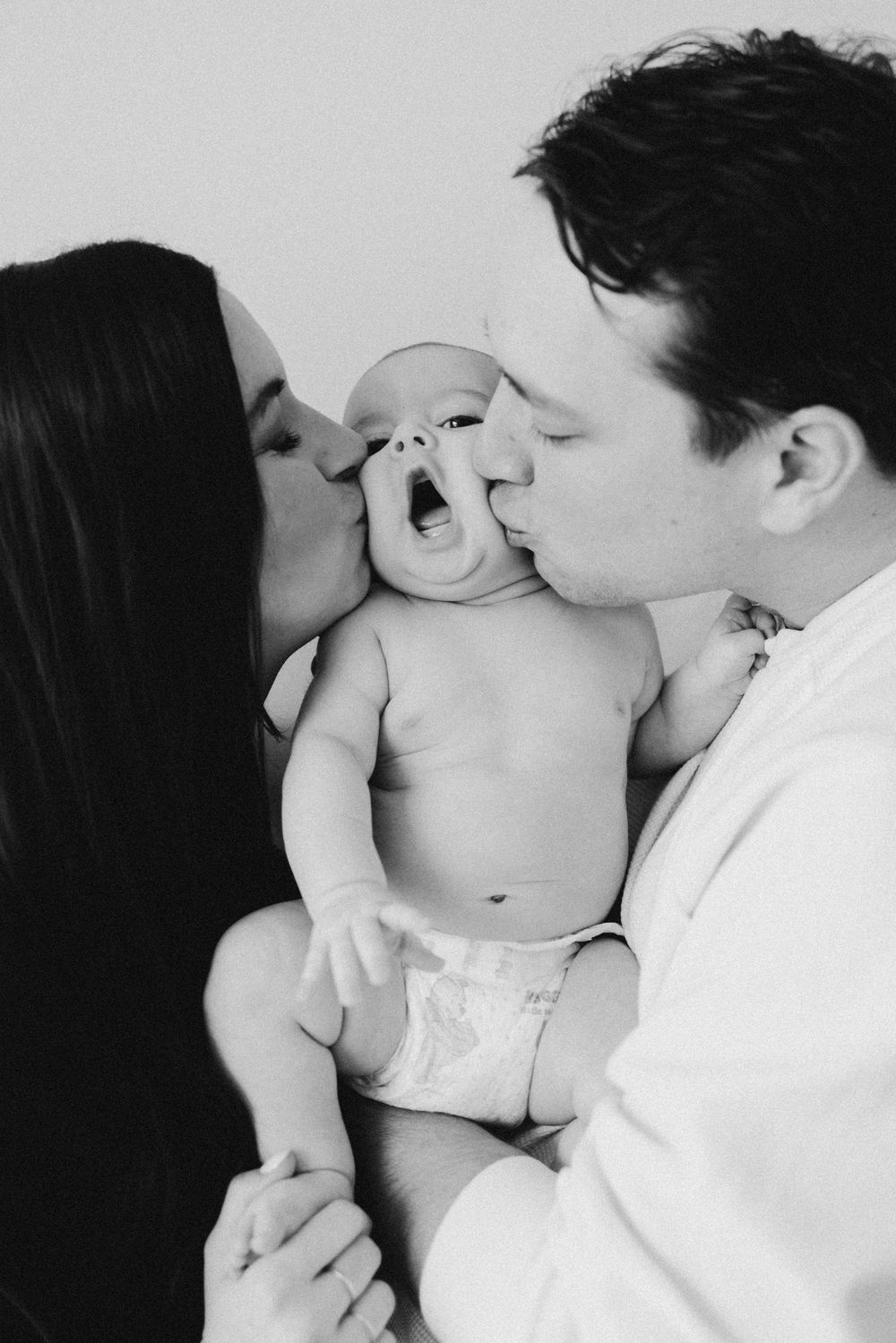 Parents sharing tender kisses with baby in black and white portrait series.