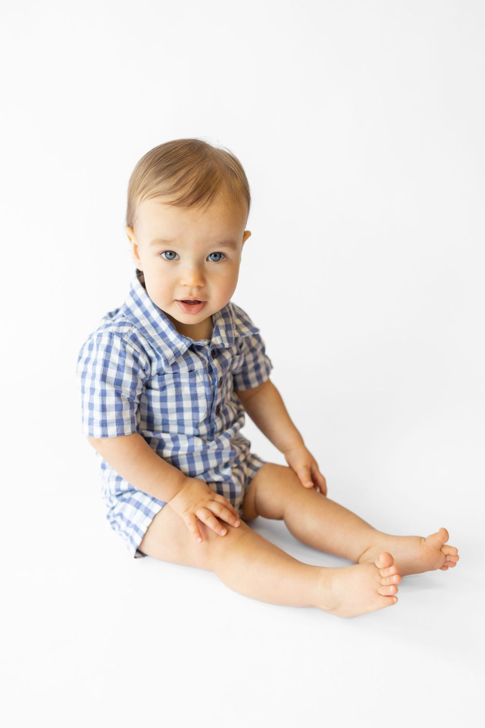 Baby wearing blue gingham outfit sits on white background showing bare feet.