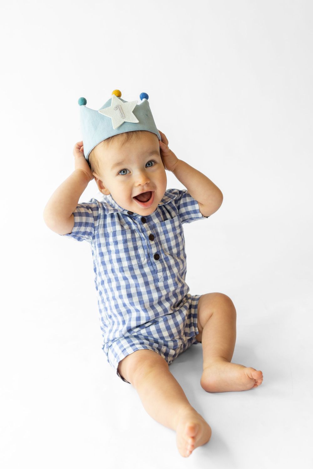 Laughing baby in blue gingham romper and cap plays on white backdrop.