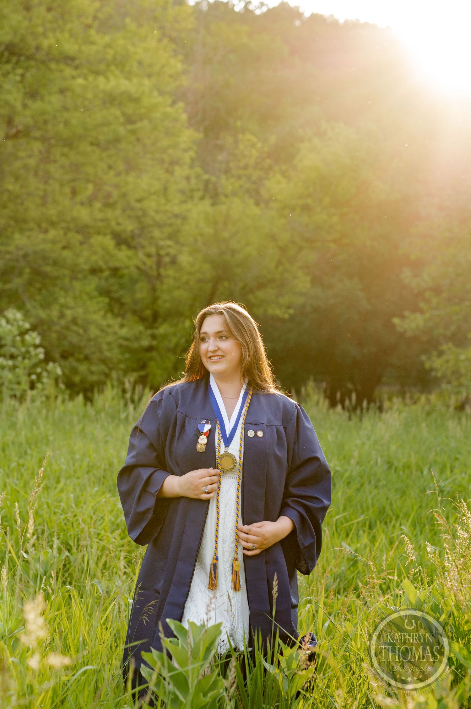 Graduate in navy blue academic regalia stands in sunlit meadow.