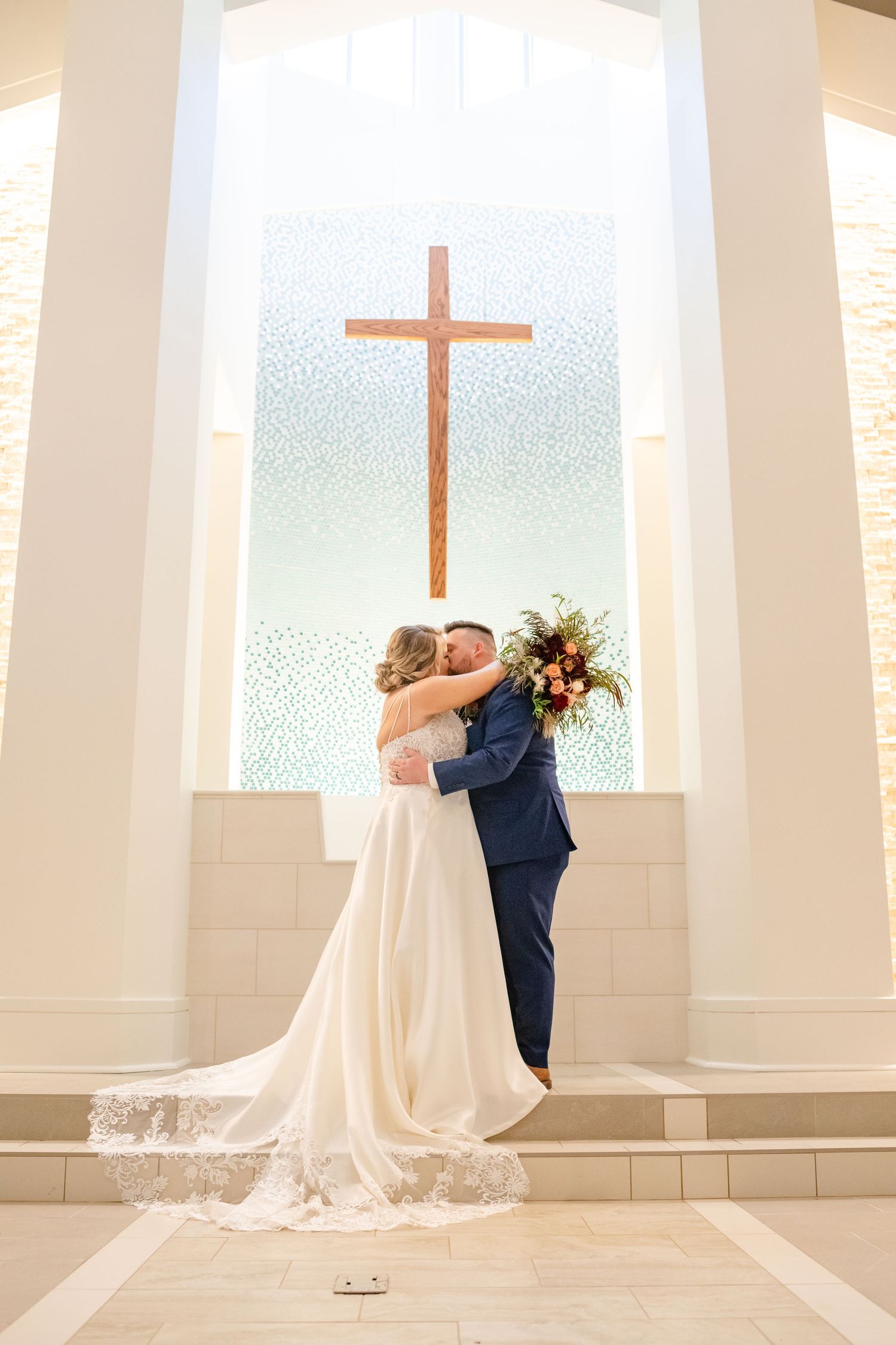 Wedding couple embracing in front of a church window with illuminated cross.