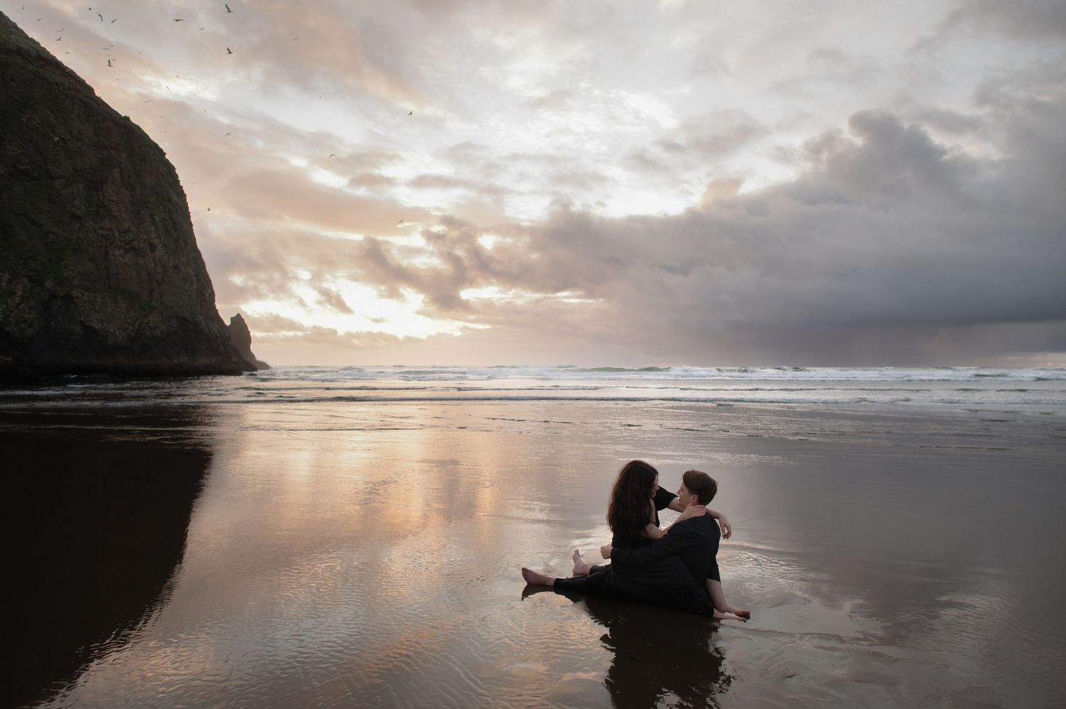 Silhouetted figures share an intimate moment on a beach at sunset with dramatic coastal cliffs.