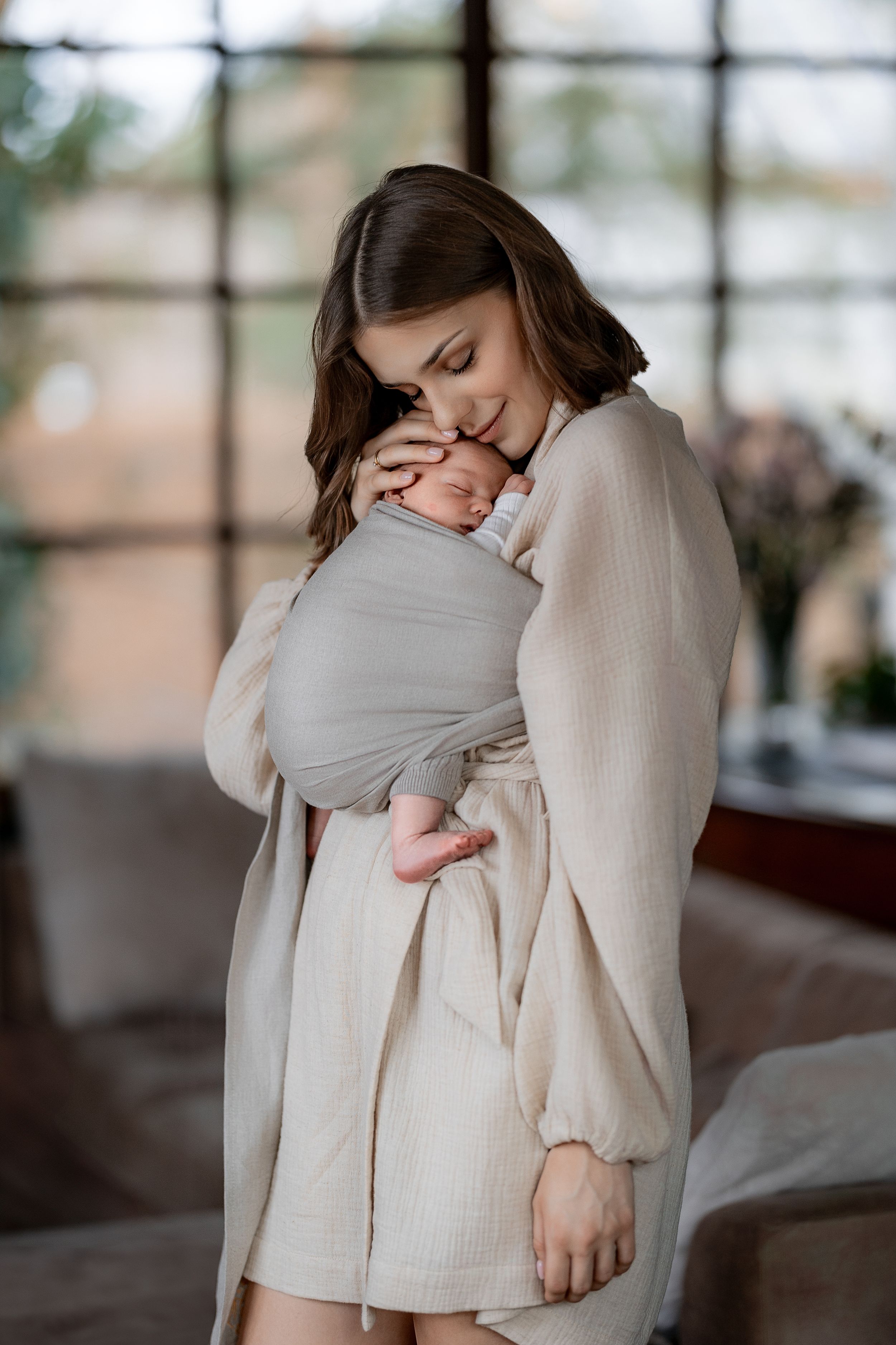 Parent tenderly holding baby wrapped in grey swaddle with soft window light.