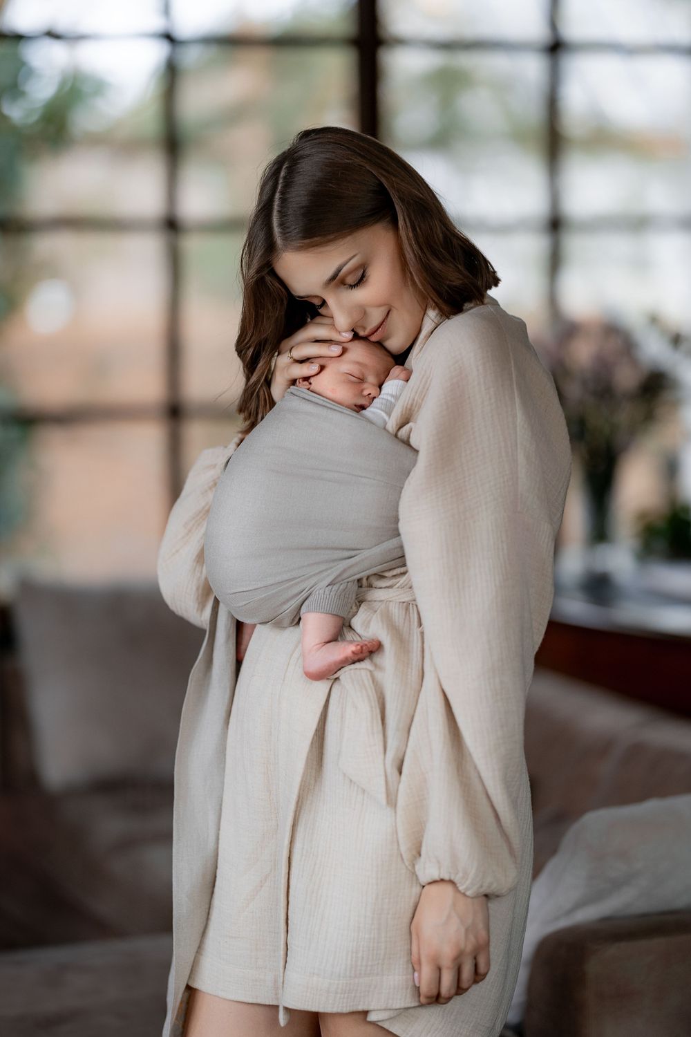 Parent tenderly holding baby wrapped in grey swaddle with soft window light.