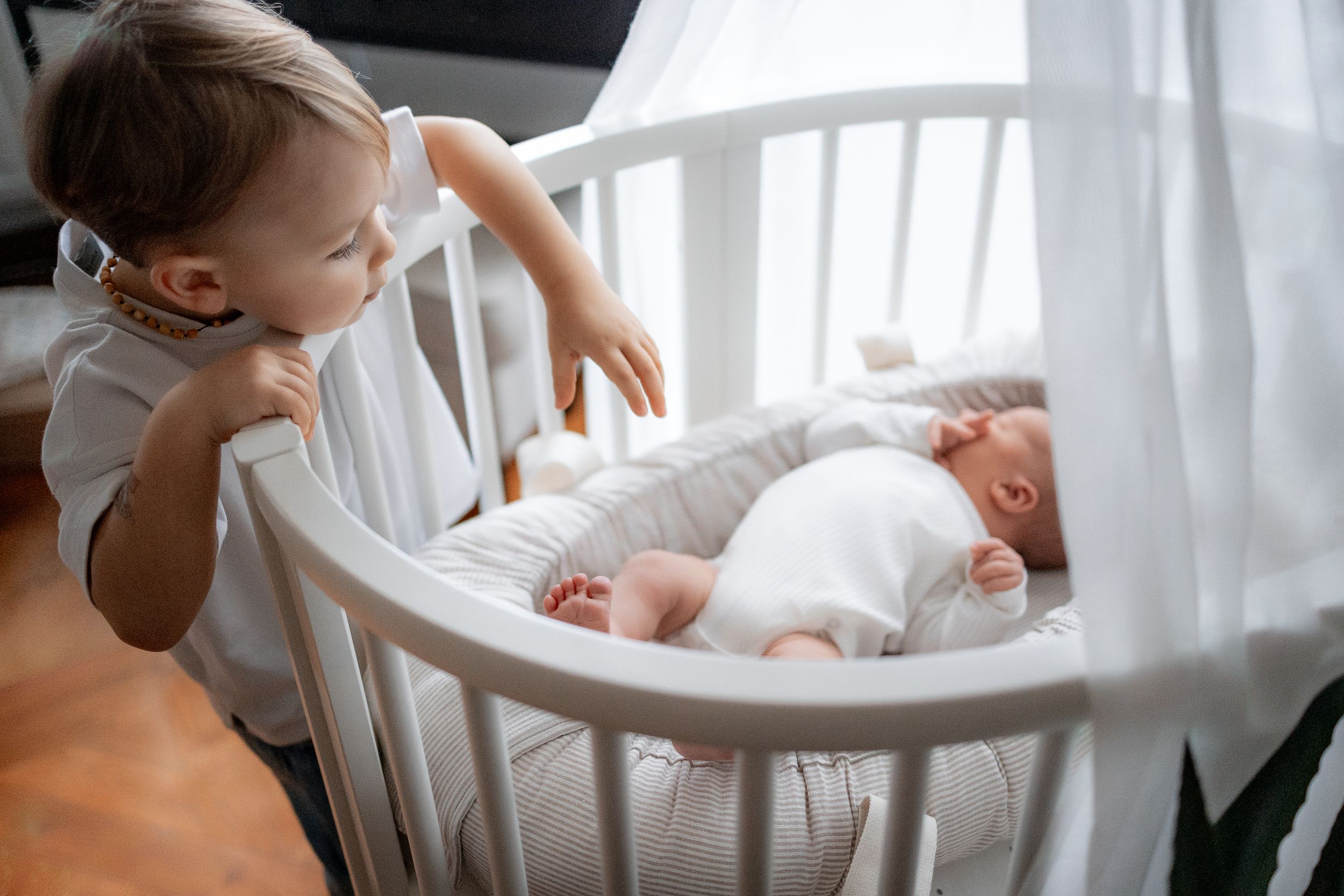 A white crib with a small baby sleeping inside while another child peers over the railing.