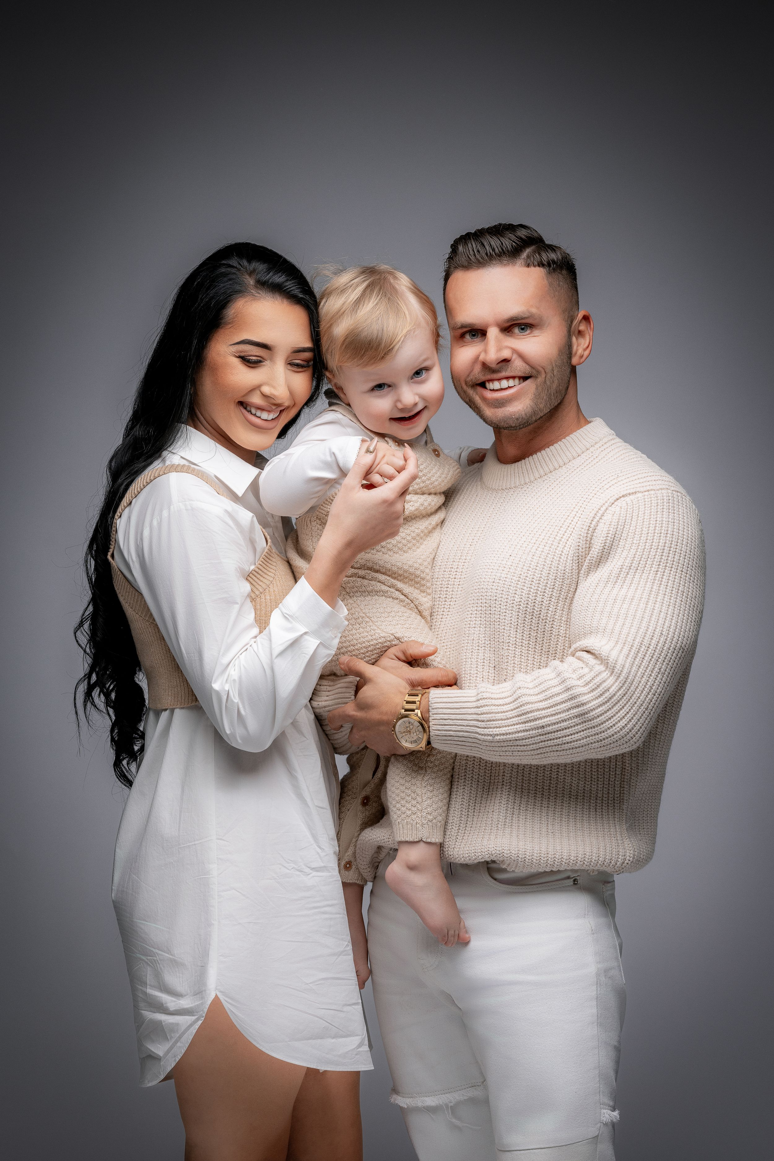 A studio portrait of a family wearing white and cream colored outfits against a grey background.