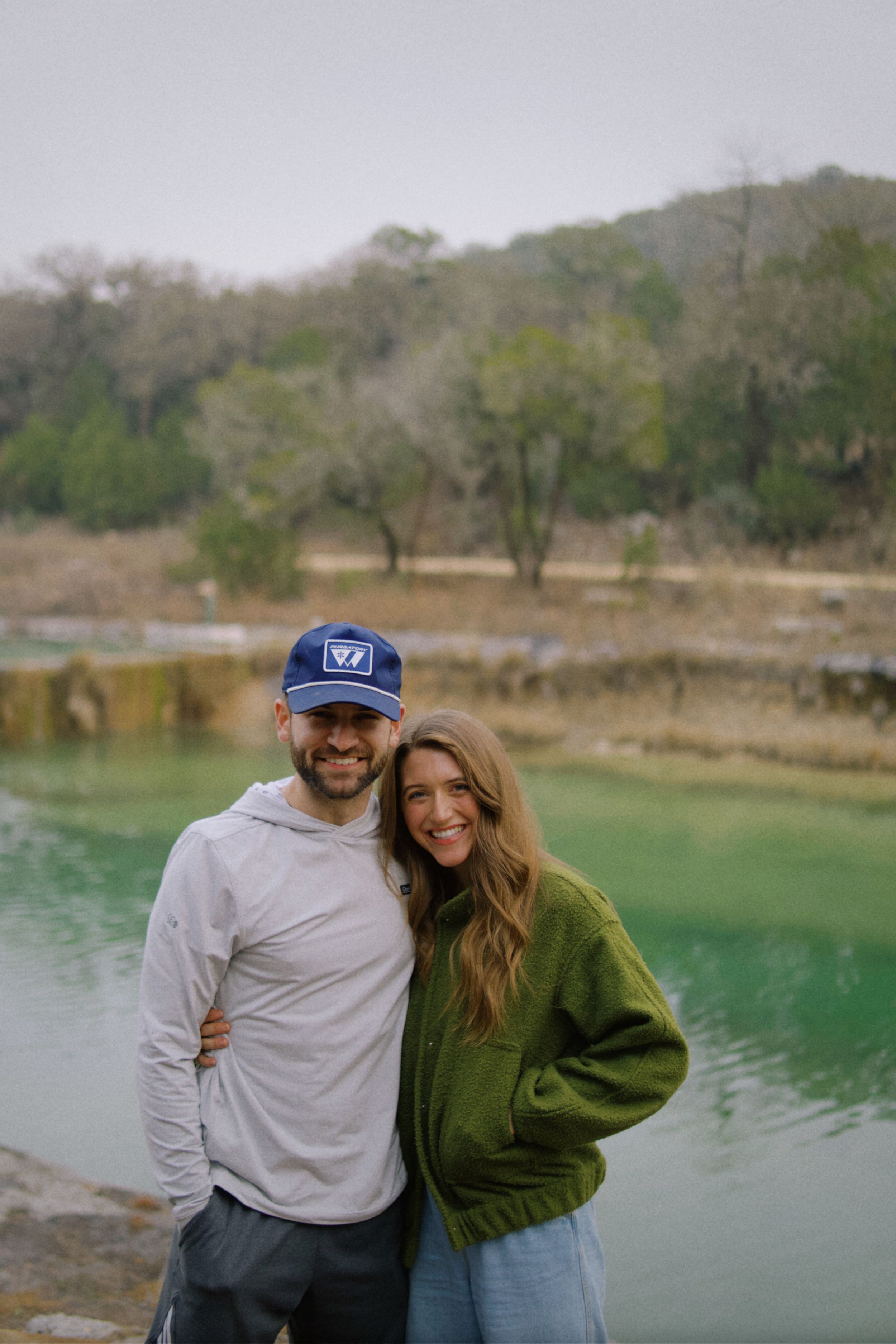 Casual couple in outdoor wear smiling together by a green lake with trees in background.
