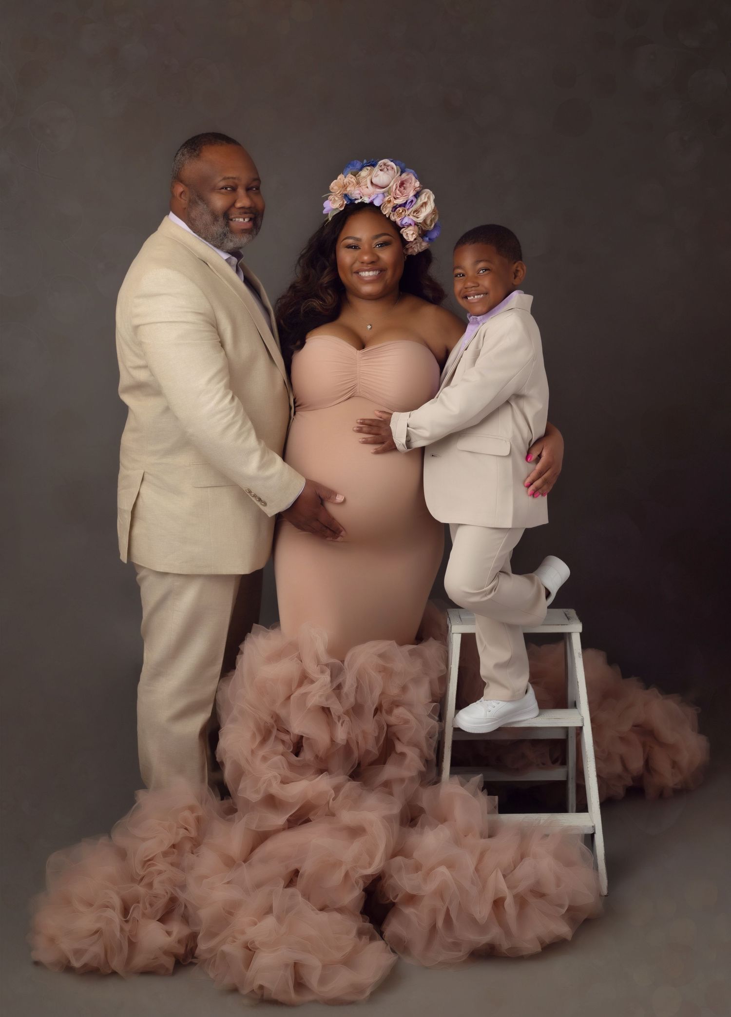 A family luxury style maternity photoshoot features a couple in beige outfits with a child on a stool against a dark backdrop.