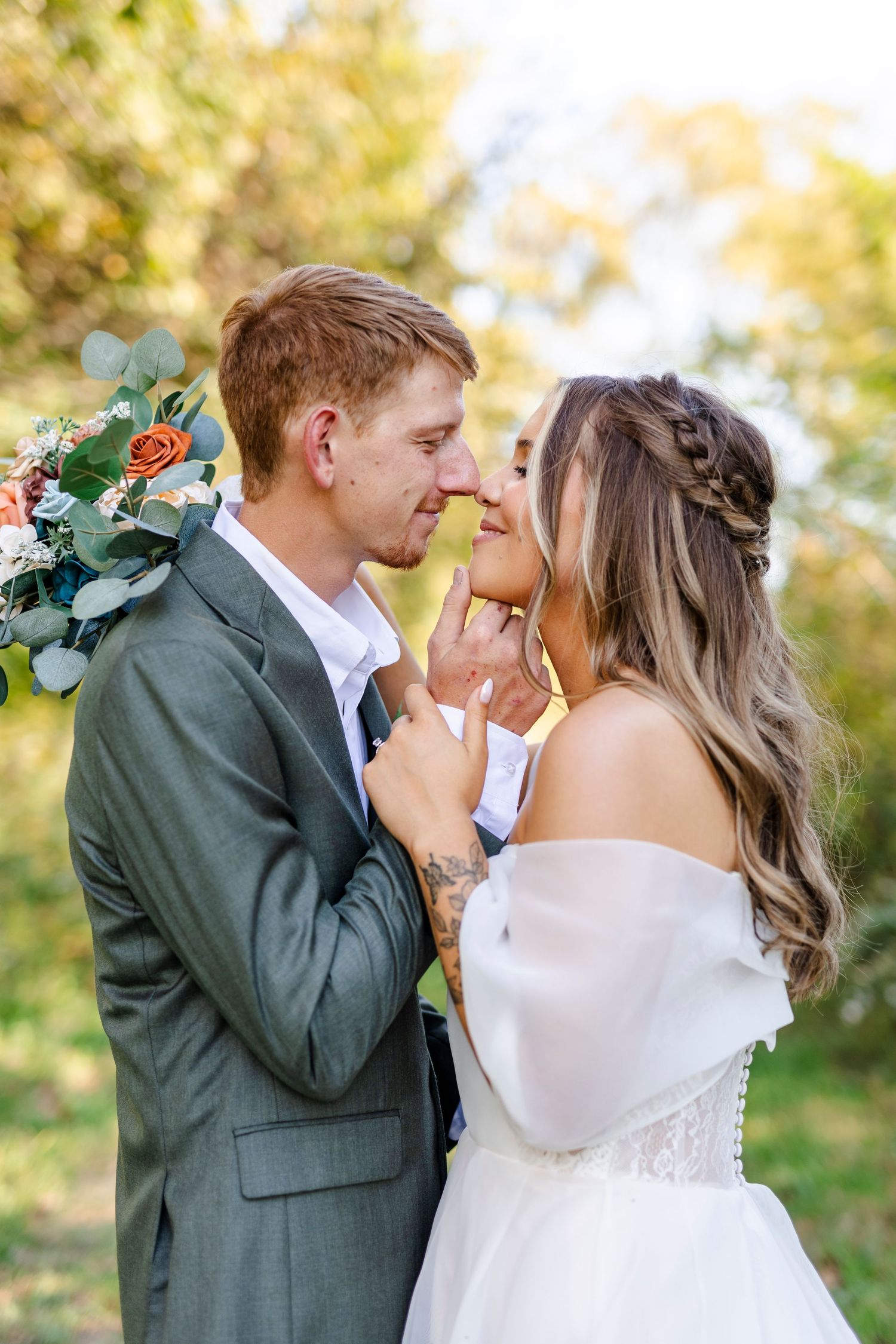 A groom in a gray suit and bride in an off-shoulder dress share a romantic moment outdoors.