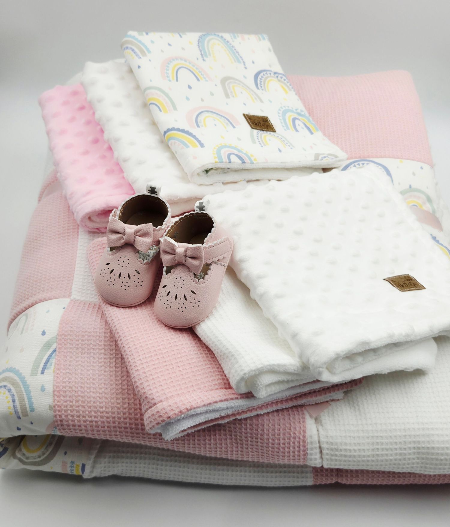 Soft pink and white baby items including blankets, booties and a rainbow print fabric arranged in a cozy display.