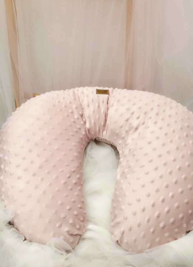 Pink minky dot nursing pillow with gold label, displayed in a soft white bassinet setting.