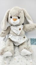 Soft plush gray bunny stuffed animal with long floppy ears wearing a floral print bib, seated on a white blanket.