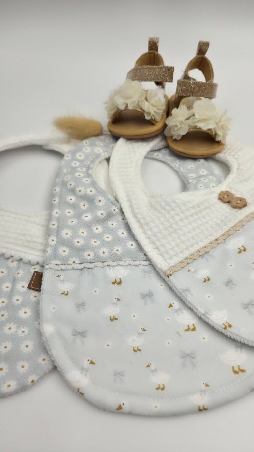 Three baby bibs in floral and waffle patterns displayed flat with a pair of gold glitter sandals with chiffon flowers on white background.