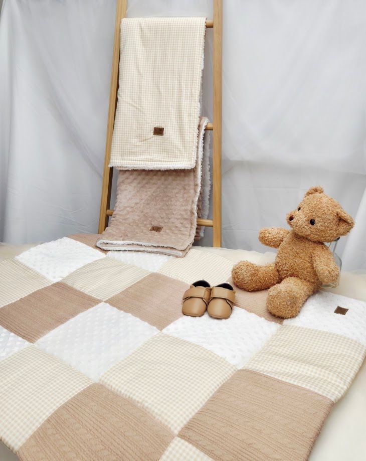 Neutral beige patchwork baby play mat with teddy bear, tiny shoes, and waffle-knit blanket on wooden ladder.