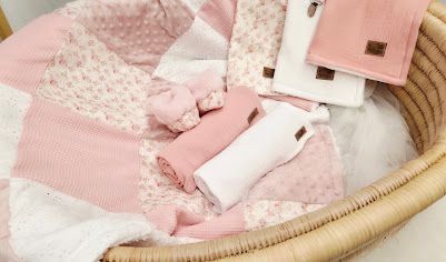 Pink baby gift set with knit blanket, muslin wrap, mittens, and velvet cloth in a wicker Moses basket.