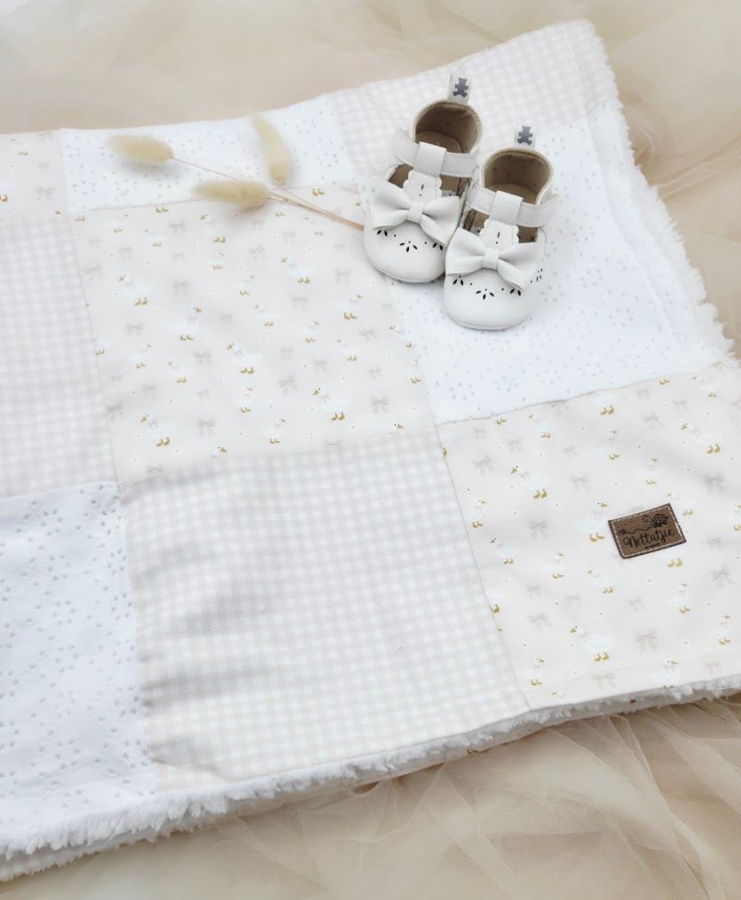 White patchwork baby blanket with eyelet lace and gingham squares, paired with white baby shoes.