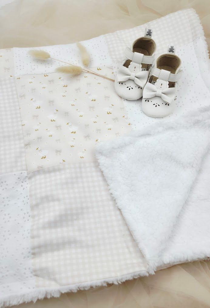 White baby blanket with patchwork fabric squares and tiny white leather bow shoes on a soft textured surface.