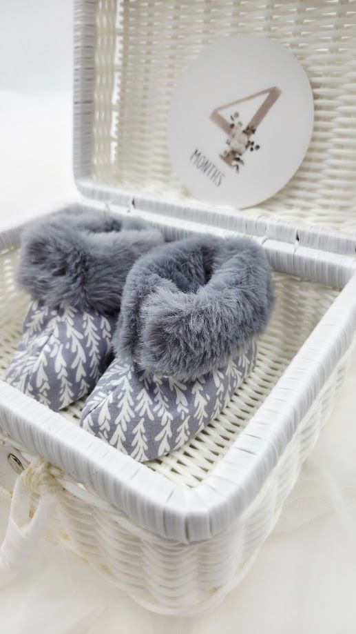 Gray fur-trimmed baby booties with white leaf pattern displayed in a white wicker basket with a 4 months milestone card.