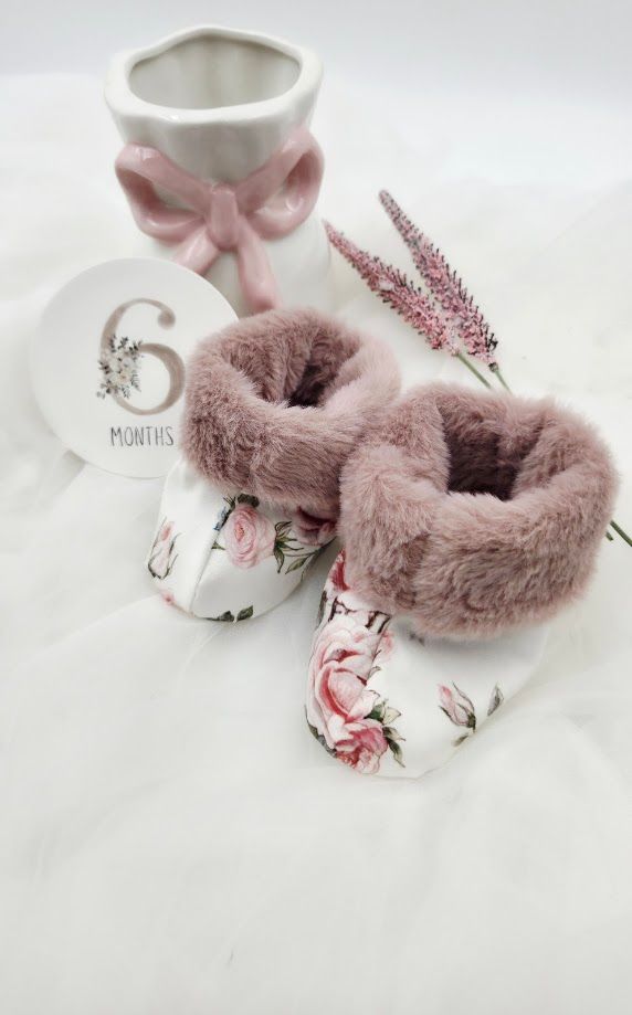 Floral baby booties with pink faux fur trim beside a 6 months milestone disc and ceramic vase.