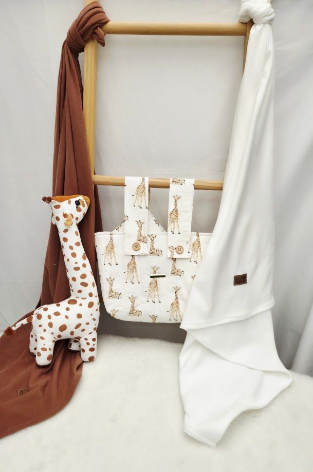 Giraffe-themed baby gift set with plush toy, white blanket, and patterned bag on wooden rack.