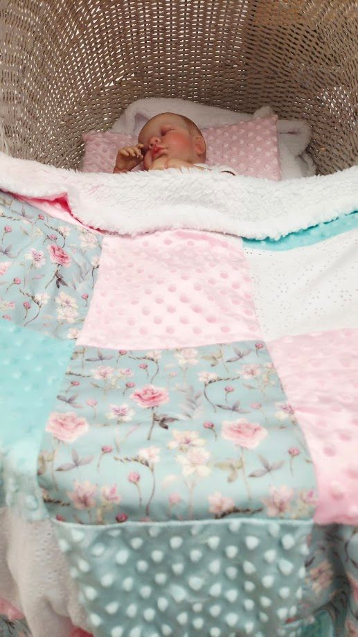 Newborn baby sleeping peacefully in a wicker bassinet covered with a colorful patchwork quilt in pink, teal, and floral patterns.