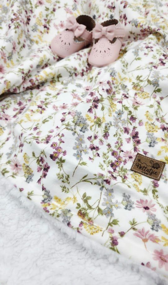 Soft floral baby blanket with pink and purple wildflowers, paired with pink bow baby shoes on a white sherpa surface.