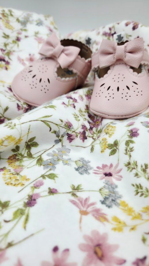 Pink leather baby shoes with bow detail on floral white fabric background.