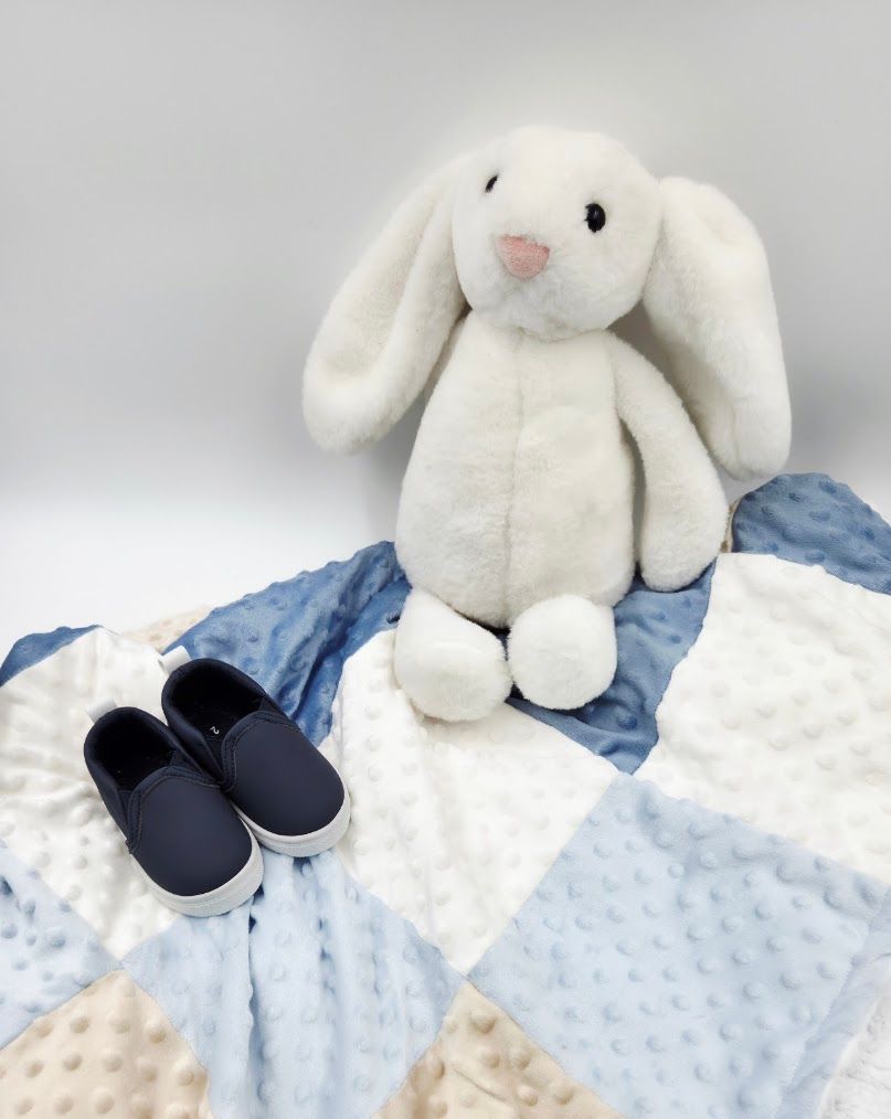 White plush bunny toy with blue and cream patchwork minky baby blanket and navy infant slip-on shoes.