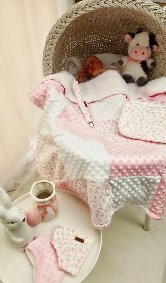 Newborn baby resting in a wicker bassinet with pink floral minky blanket, cow plush toy, and baby accessories on a side table.