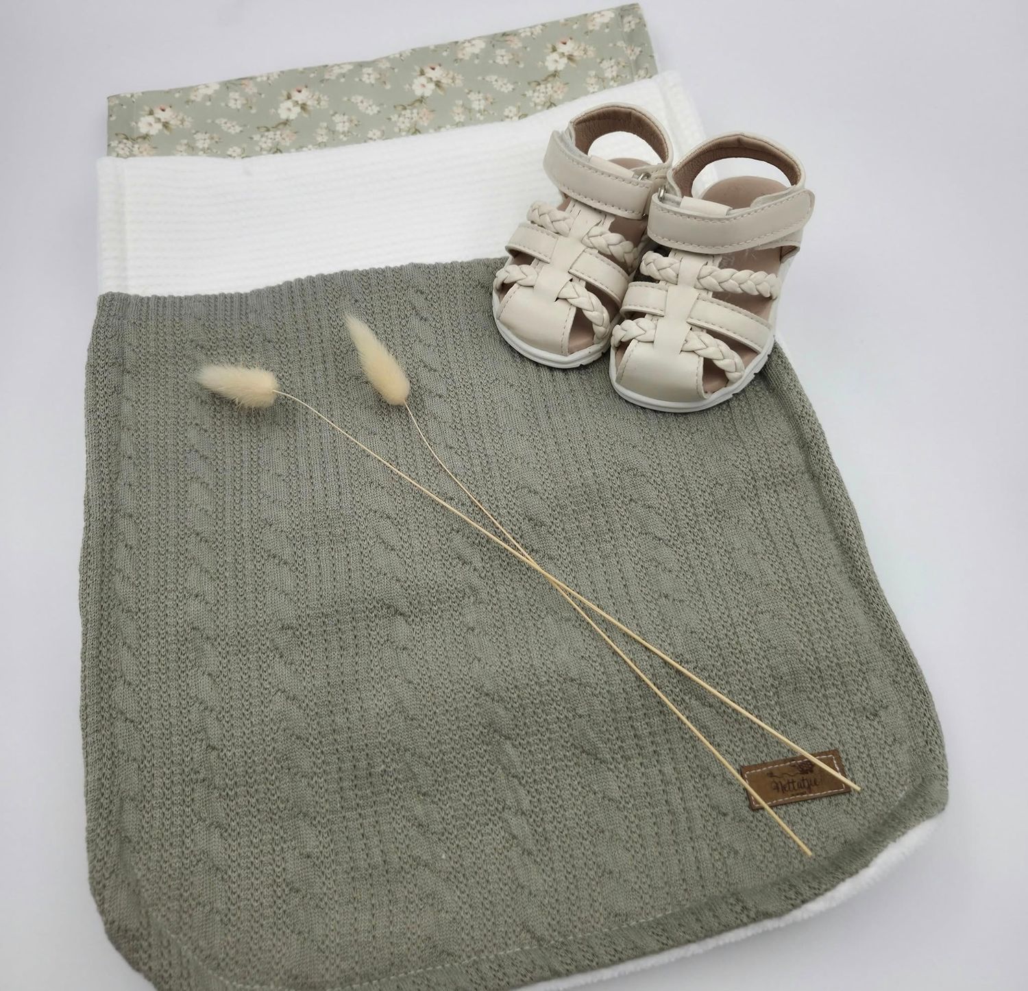 Gray knit baby blanket with dried wheat stems and beige sandals on white background.