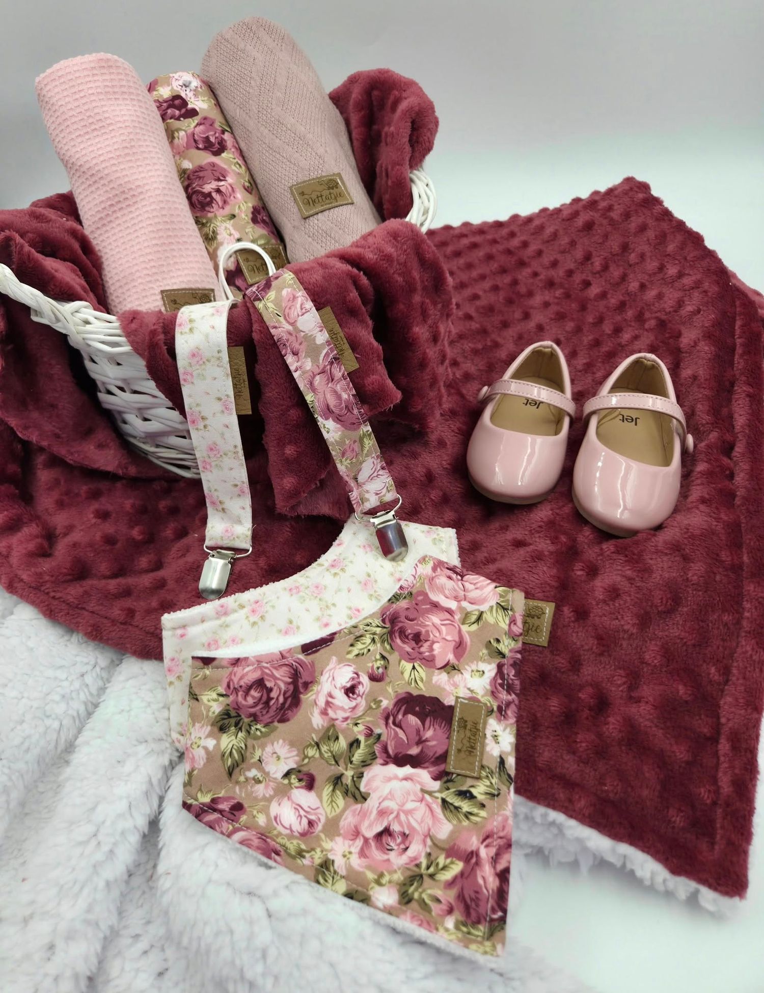 Burgundy minky blanket with floral print baby accessories and pink shoes in white wicker basket.