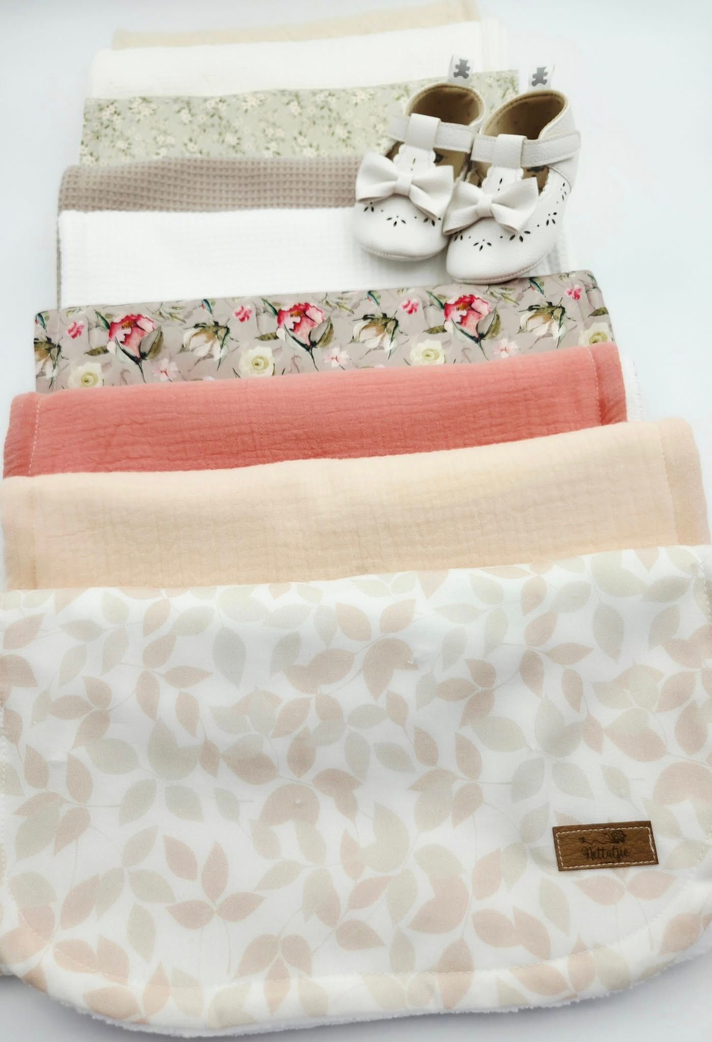 Layered baby blankets in neutral tones with floral and leaf patterns and white leather shoes.