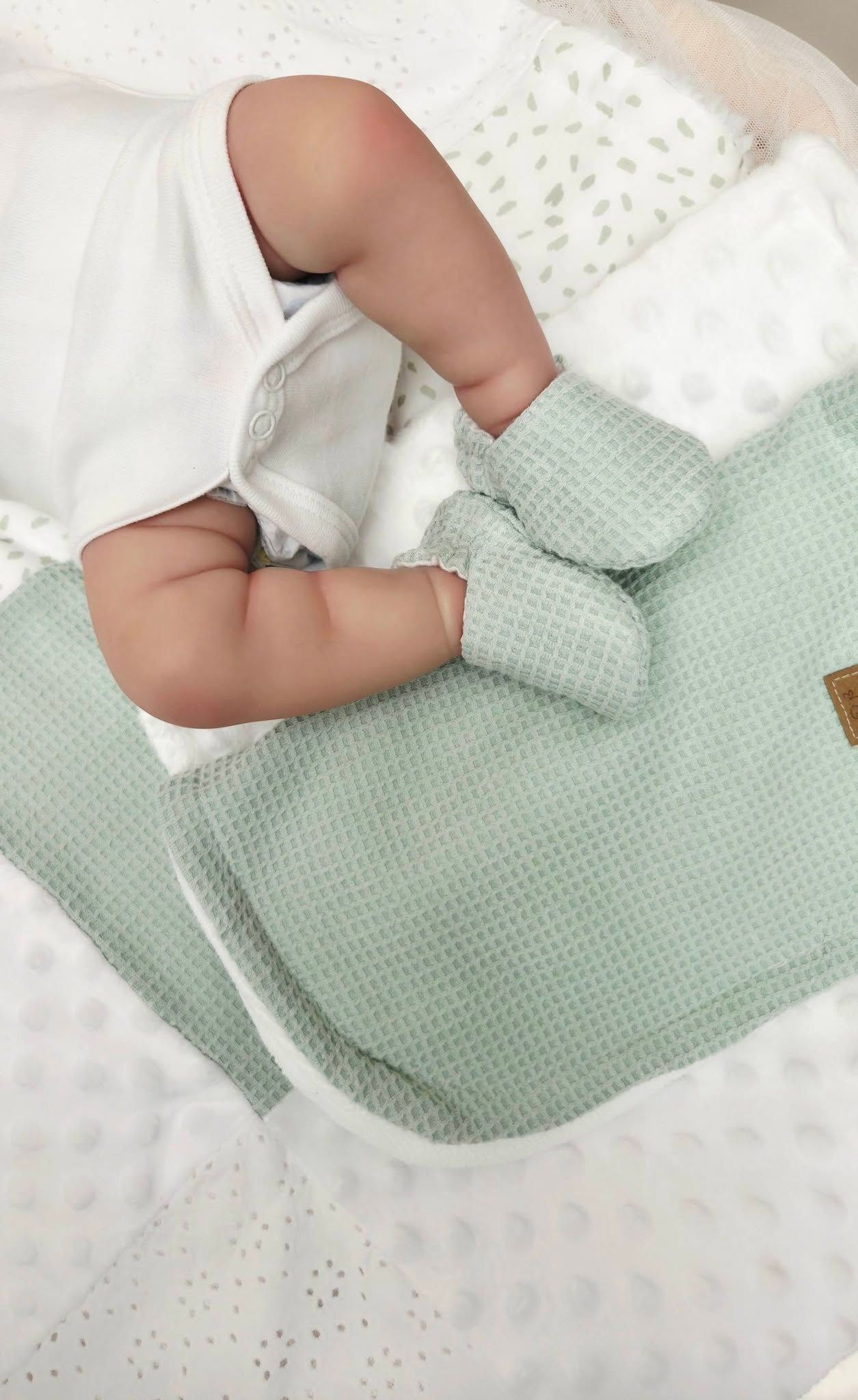Newborn baby resting on a soft sage green waffle knit blanket with matching mittens on white bedding.