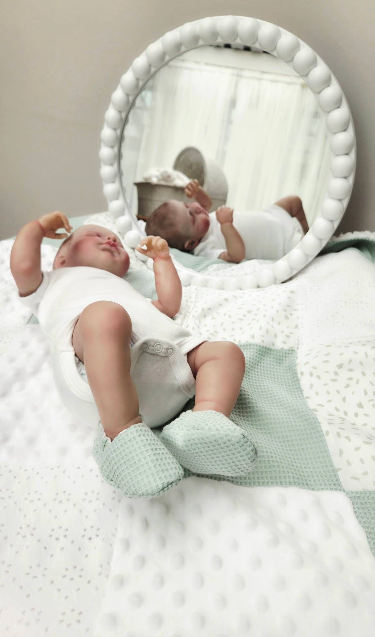 Newborn baby in white onesie and mint booties lying on white blanket beside round mirror reflecting their peaceful sleeping pose.