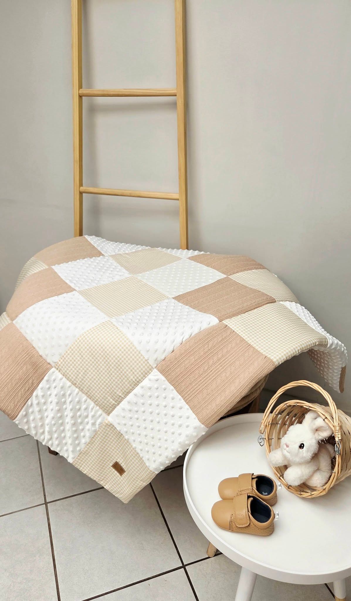Beige and white patchwork baby blanket draped over a ladder with a small white table holding baby shoes and a toy.